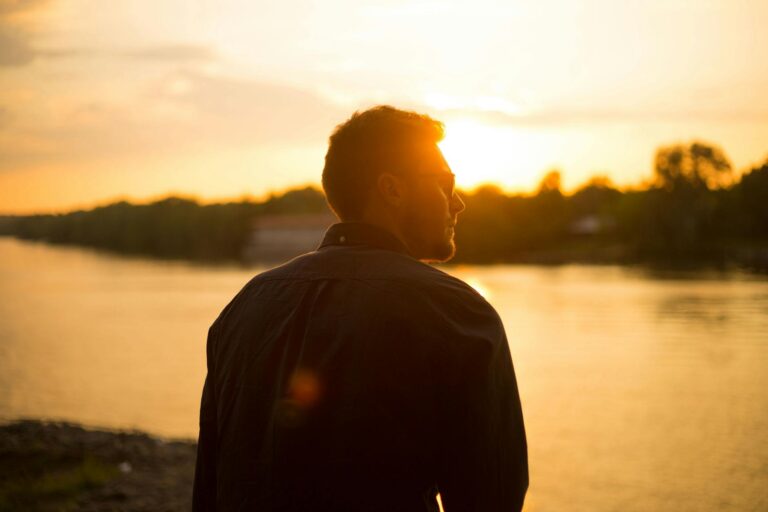 man sitting near large body of water under clear sky during sunset